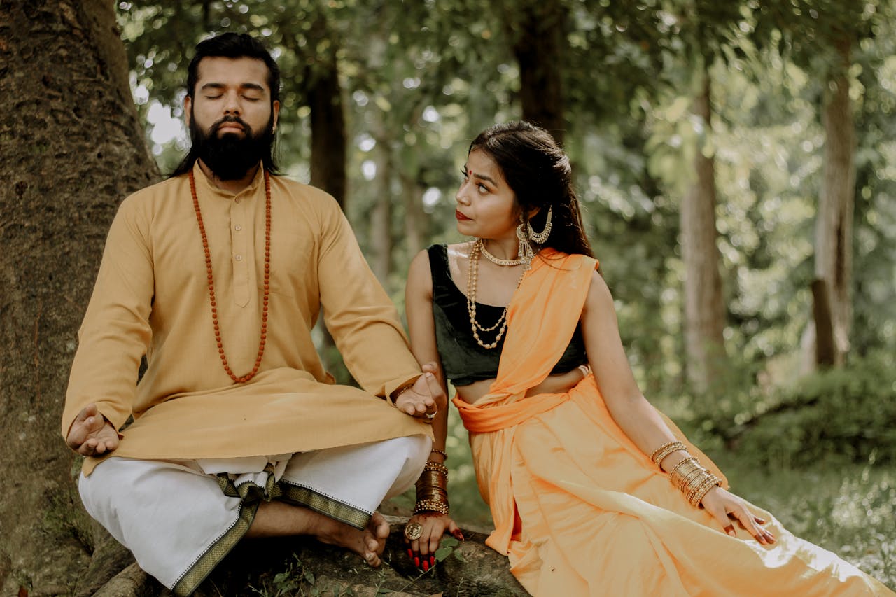 gallery-4 A couple in traditional attire meditating and sitting together in a lush green forest.